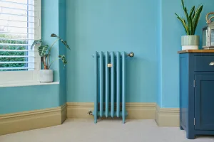 Tall Emmeline III cast iron radiator in a teal-blue finish on feet, with valves, against a blue wall beside a shuttered window and blue cabinet.