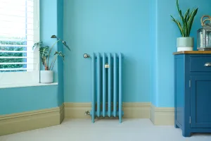 Emmeline III 870mm cast iron radiator painted blue, on feet beside a window, with a thermostatic valve and a blue cabinet on the right.