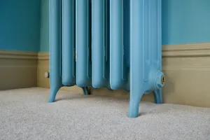 Low view of the feet of an Emmeline III cast iron radiator painted light blue, standing on beige carpet against a teal wall and cream skirting.