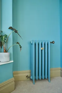 Plain six-section Emmeline III cast iron radiator on feet, painted blue against a matching blue wall, with brass valves and a beige carpet.
