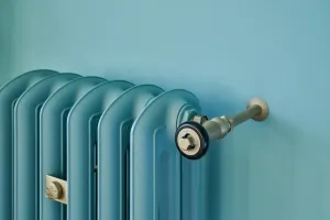 Close-up of the top of an Emmeline III cast iron radiator painted teal against a matching wall, with a brass-toned valve and wall stay.