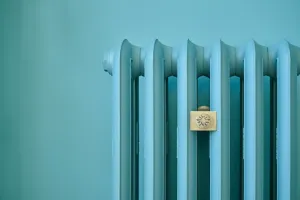 Close-up of the top of a plain Emmeline III cast iron radiator painted light blue, with a brass bee-emblem plate, against a matching blue wall.