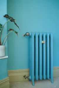 Emmeline III 870mm cast iron radiator on feet in matching blue against a blue wall, with brass valves, olive skirting and a potted plant.