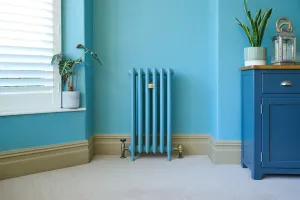 Emmeline III 870mm cast iron radiator on feet, painted blue to match the wall, with brass angled valves, beside a shuttered window.