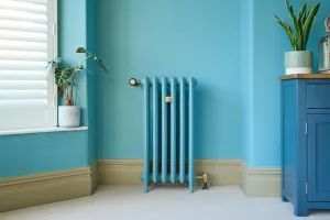 A slim six-section Emmeline III cast iron radiator in a blue finish on feet, with brass valves, against a turquoise wall beside a window.