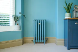 Emmeline III 870mm cast iron radiator painted blue to match the wall, on feet with valves, beside a shuttered window and a blue cabinet.