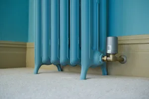 Lower half of a blue Emmeline III cast iron radiator on feet, with a brass angled valve and smart thermostatic head, on cream carpet.