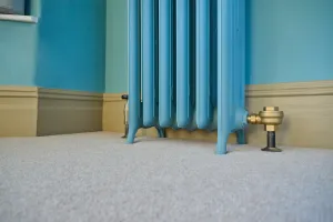Low view of the feet of a light blue Emmeline III cast iron radiator on beige carpet, with a brass valve, cream skirting and teal walls.