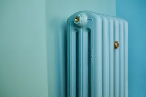 Top corner of an Emmeline III cast iron column radiator in a pale blue finish with brass end plugs, set against turquoise walls.