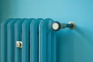 Top corner of an Emmeline III cast iron radiator painted teal blue against a matching wall, with a satin brass valve, pipe and small badge.