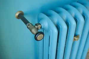 Close-up of a light blue Emmeline III cast iron radiator with an aged brass angled valve and wall stay against a teal painted wall.