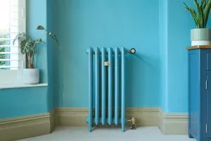Emmeline III 870mm cast iron radiator in blue on feet against a turquoise wall, with brass valves, beside a window and blue cabinet.