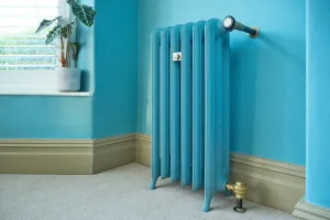 Emmeline III 870mm cast iron radiator on feet in a blue finish, with brass valve and wall stay, against a turquoise wall beside a window.