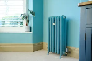 A tall, slim Emmeline III cast iron radiator on feet in a blue finish, with a brass valve and smart head, beside a window on a cream carpet.