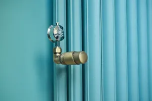Close-up of a brass thermostatic valve with a chrome air vent on a plain teal-blue Emmeline III 870mm cast iron radiator against a matching wall.