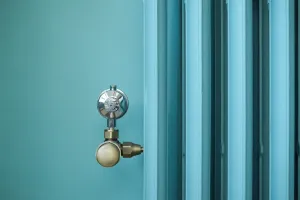 Close-up of a pale blue plain-column Emmeline III cast iron radiator with a brass angled valve and polished chrome head against a teal wall.