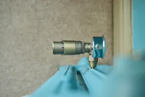 Close-up of a thermostatic radiator valve in an antique brass-style finish on top of a light blue Emmeline III cast iron radiator, beige wall behind.