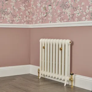 Emmeline I cast iron radiator in Chalk on feet, with brass valves, against a pink wall below blossom wallpaper on a herringbone floor.