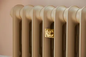 Close-up of an Emmeline cast iron radiator in Library Truffle with a polished brass square valve cap, against a pale pink wall.
