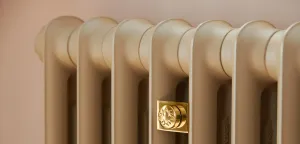 Close-up of a plain cast iron column radiator in a matt Truffle finish with a square polished brass wall stay, against a pale pink wall.