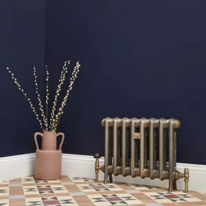 Emmeline I cast iron radiator in Beaten Brass on feet with brass valves, against a navy wall above a patterned tiled floor beside a pink vase.