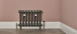 Emmeline I 465mm cast iron radiator in Black Iron with wall stays and nickel valves, against a pink wall above a white skirting and herringbone floor.