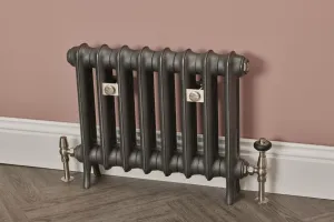 Seven-column Emmeline I cast iron radiator in Black Iron on feet, with nickel valves and wall stays, against a pink wall above herringbone flooring.