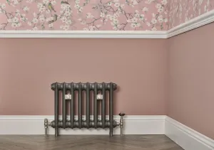 Emmeline I 465mm cast iron radiator in Black Iron on feet with nickel valves, against a pink wall below floral wallpaper and a dado rail.
