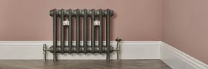 Emmeline I cast iron radiator in Black Iron on feet against a pink wall, with nickel valves, wall stays and a white skirting board.