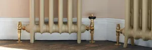 Bases of two cream-painted Emmeline cast iron radiators on feet in a bay, with Unlacquered Brass valves, white skirting and a wooden floor.