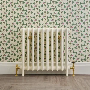Emmeline I 665mm cast iron radiator in cream on feet, with brass valves and wall stays, against clover wallpaper above a wood floor.
