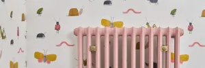 Emmeline I 665mm cast iron radiator in Little Greene Blush 267 with brass wall stays, against white wallpaper printed with insects.