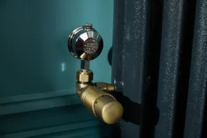 Close-up of an antique brass thermostatic valve with a chrome air vent on the side of an Emmeline I radiator in Midnight Ink, against a teal wall.