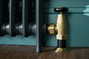 Close-up of an Emmeline I cast iron radiator foot in Midnight Ink with a brass angled valve, black wheel head, against a teal skirting on a wooden floor.