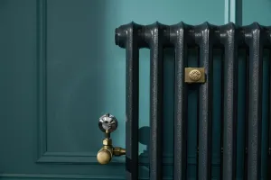 Close-up of an Emmeline I cast iron radiator in glossy Midnight Ink against a teal panelled wall, with a brass valve and a brass bee-motif wall stay.
