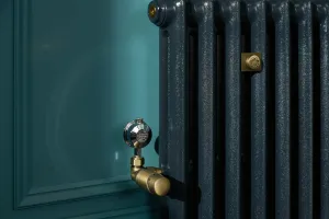 Close-up of an Emmeline I cast iron radiator in Midnight Ink with a brass angled valve, brass wall stay and chrome lockshield against a teal panelled wall.