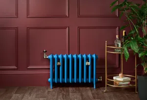 Bright blue Emmeline II cast iron radiator on feet with brass valves and wall stays, against a panelled burgundy wall beside a brass drinks trolley.