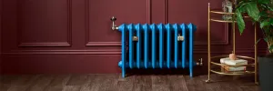 Emmeline II 470mm cast iron radiator in bright blue on feet, with brass valves and wall stays, against a burgundy panelled wall beside a brass trolley.