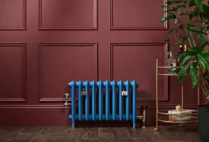 A blue Emmeline II 470mm cast iron radiator on feet against a burgundy panelled wall, with angled valves, wall stays and a brass drinks trolley.