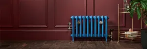 Blue Emmeline II cast iron radiator on feet against a burgundy panelled wall, with nickel valves, wall stays and a brass drinks trolley.