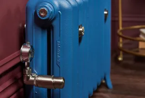Close-up of a blue Emmeline II cast iron radiator on feet with a polished nickel thermostatic valve, against a deep red panelled wall.