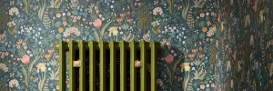 Top of an Emmeline II cast iron radiator in olive green Farrow & Ball Bancha No. 298 with two brass wall stays, against dark floral wallpaper.