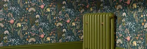 Emmeline II cast iron column radiator in Farrow & Ball Bancha No. 298, beside a matching green skirting board and dark floral wallpaper.