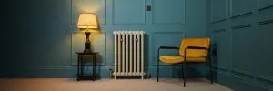 Emmeline II cast iron radiator in Portland Stone on feet against a teal panelled wall, with lamp on side table and mustard armchair.