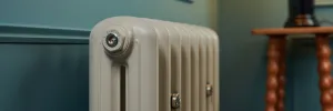 Close-up of a plain Emmeline II cast iron radiator in Little Greene Portland Stone 77 against a teal wall, beside a turned wooden side table.