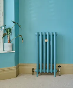 Six-column Emmeline III cast iron radiator on feet in a blue finish, with brass valves, against a blue wall beside a potted plant on a windowsill.