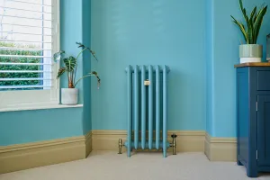 Emmeline III cast iron radiator on feet, painted turquoise to match the wall, with brass valves, beside a shuttered window on cream carpet.