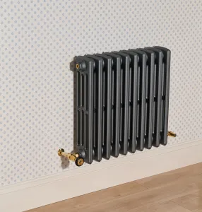 Grace 4 Column 660mm cast iron radiator in dark grey with Natural Brass valves, wall-hung above a skirting board on patterned wallpaper.
