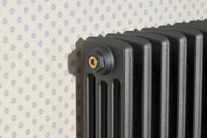 Close-up of a plain Grace four-column cast iron radiator in dark grey Pencil with a Natural Brass bleed plug, against blue star-patterned wallpaper.