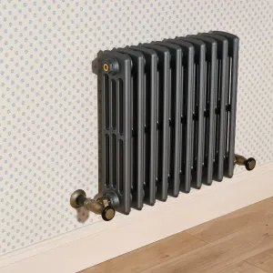 Wall-hung Grace 4 Column cast iron radiator in dark grey with Natural Brass angled valves, against star-print wallpaper above a wooden floor.
